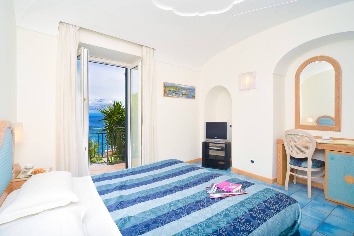 A hotel room with a bed, a desk, a television, and a balcony with a view of the sea.