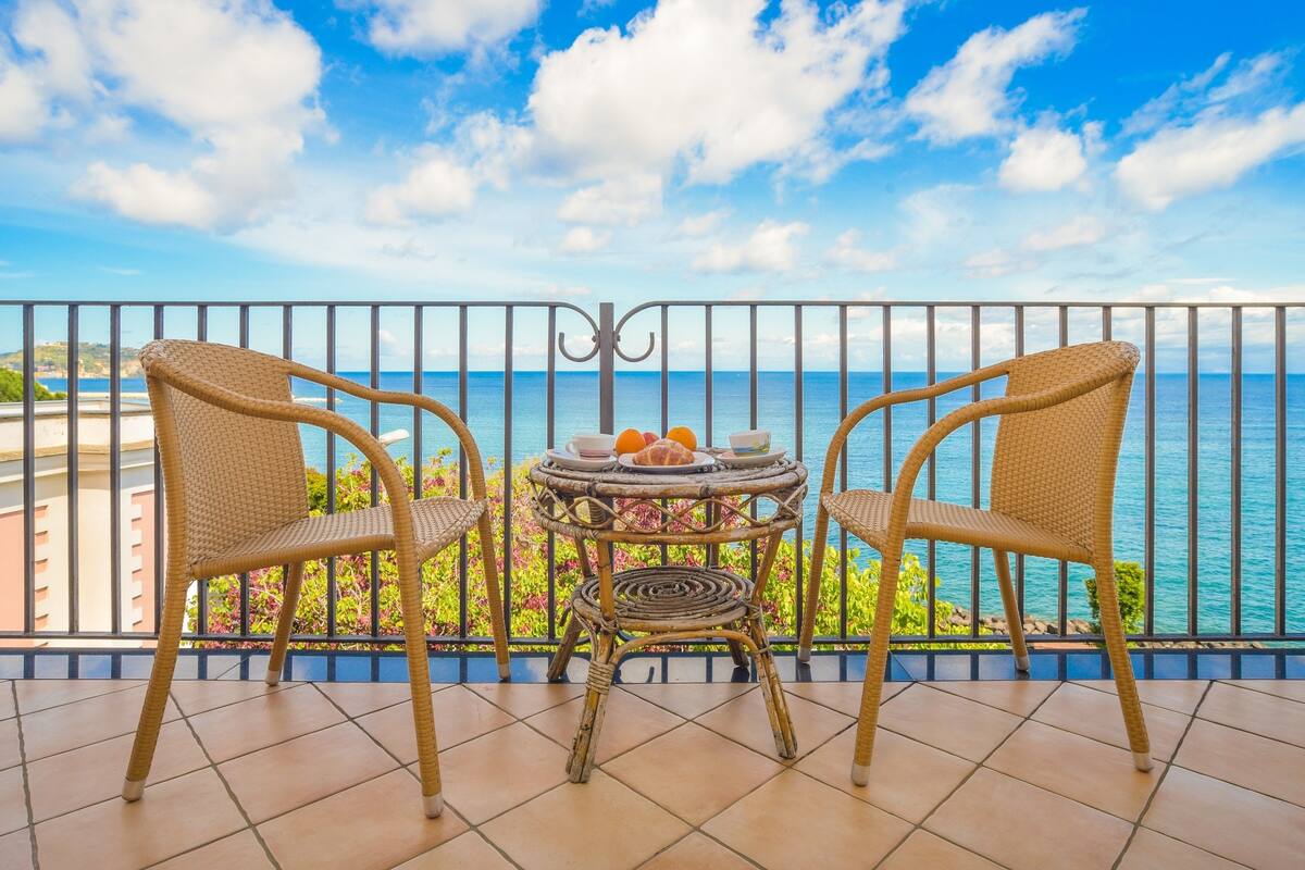 A balcony with wicker chairs and a small table, overlooking a scenic ocean view.