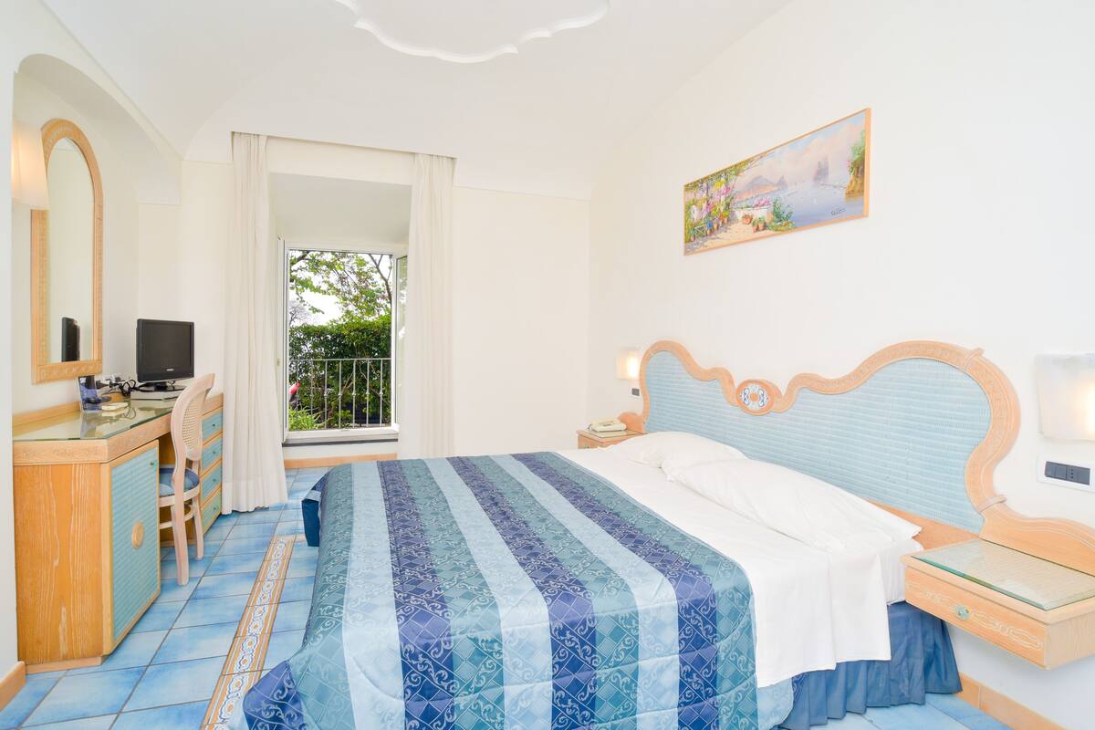 A bedroom with a bed, a desk, a chair, a nightstand, a painting, and a view of the outdoors.