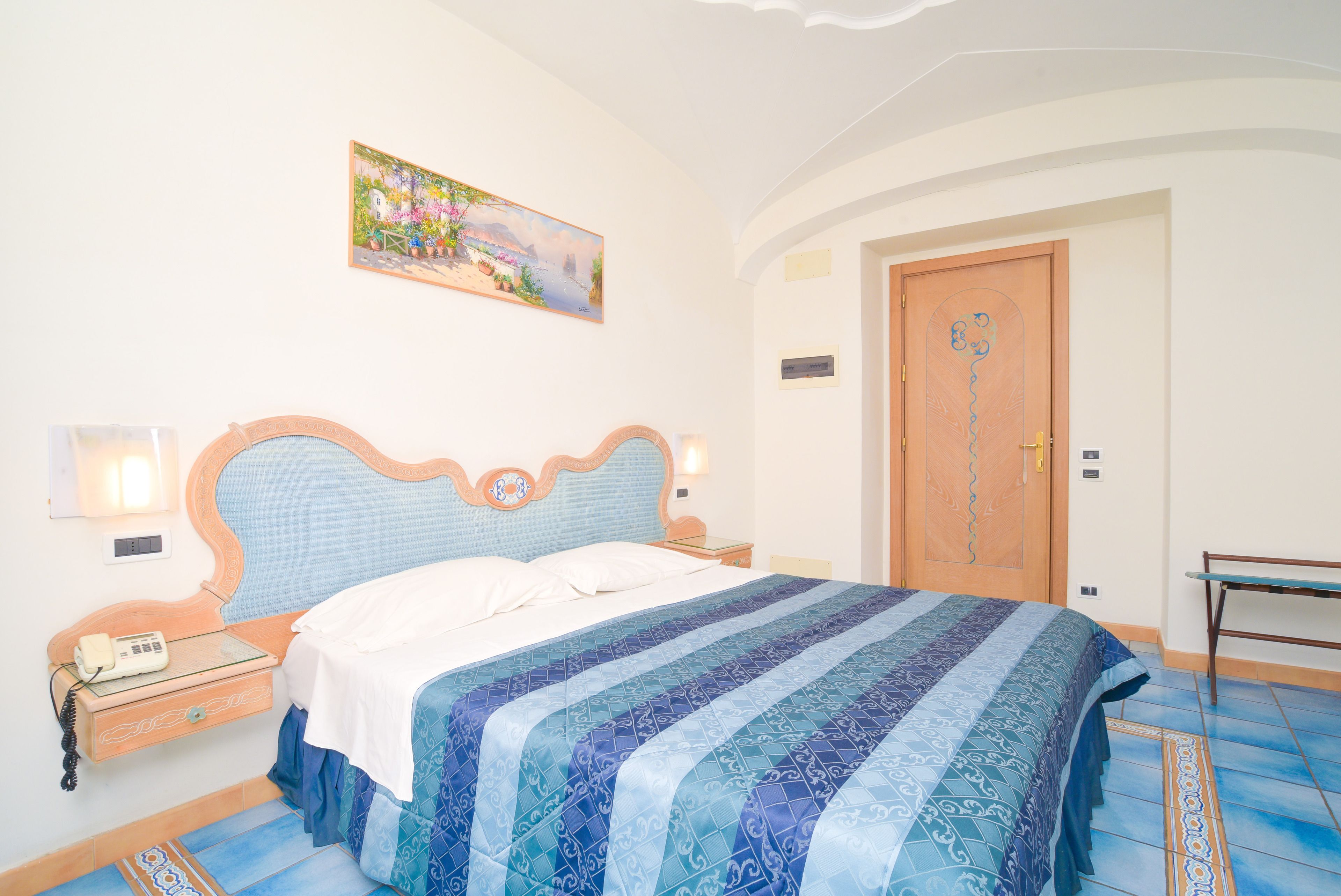 A bedroom with a bed, a nightstand, a telephone, a painting on the wall, and a door.