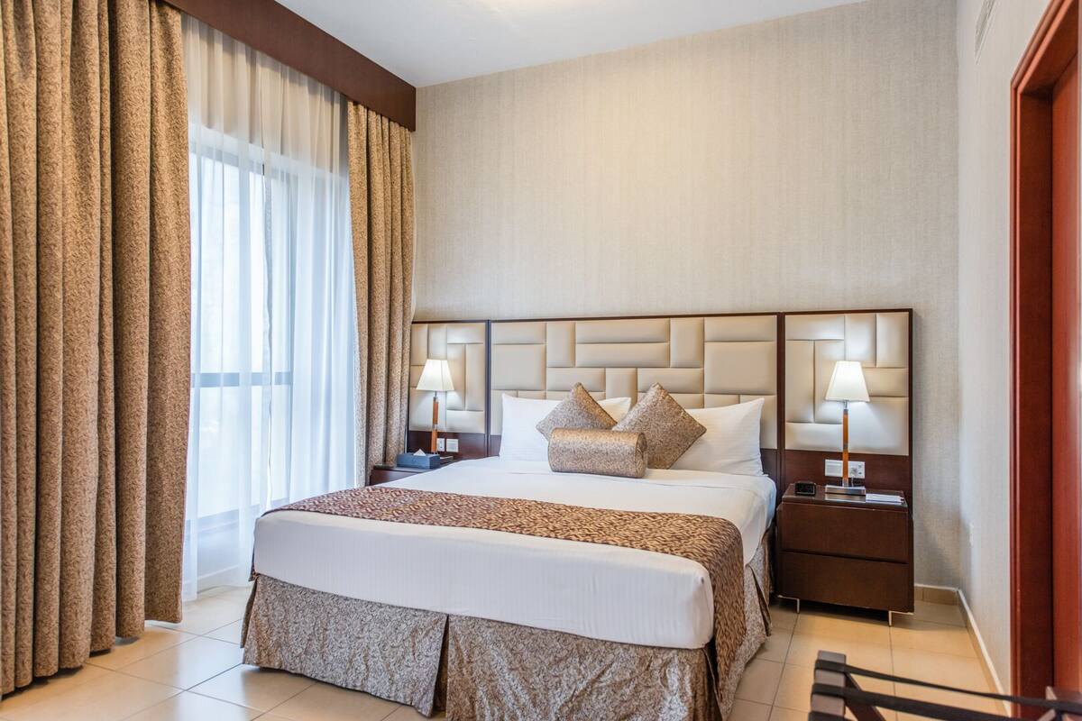 Hotelkamer met een groot bed, twee nachtkastjes met lampen, een hoofdbord met een gestoffeerd ontwerp en een raam met gordijnen.