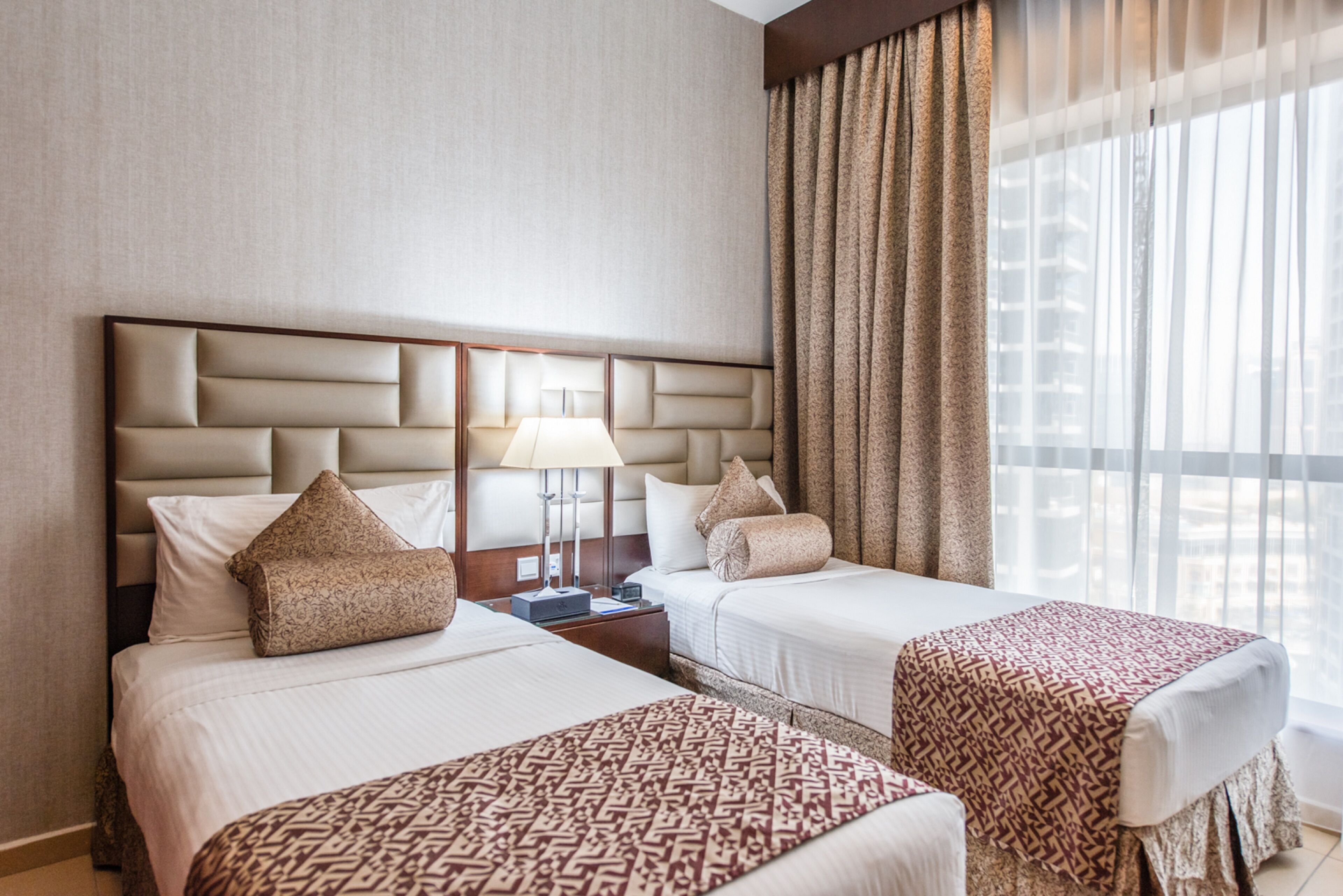 A hotel room with two beds, a large window with curtains, a nightstand with a lamp, and a view of the city.
