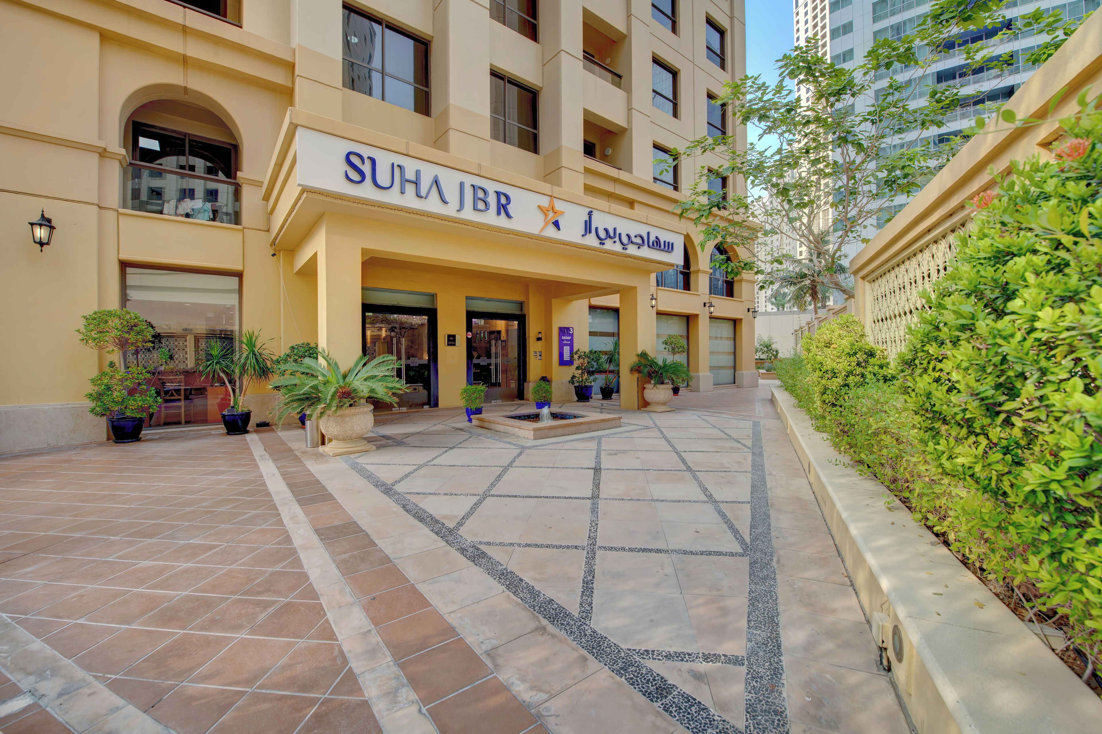 A hotel entrance with a tiled courtyard, potted plants, and a sign reading "SUHAJBR".