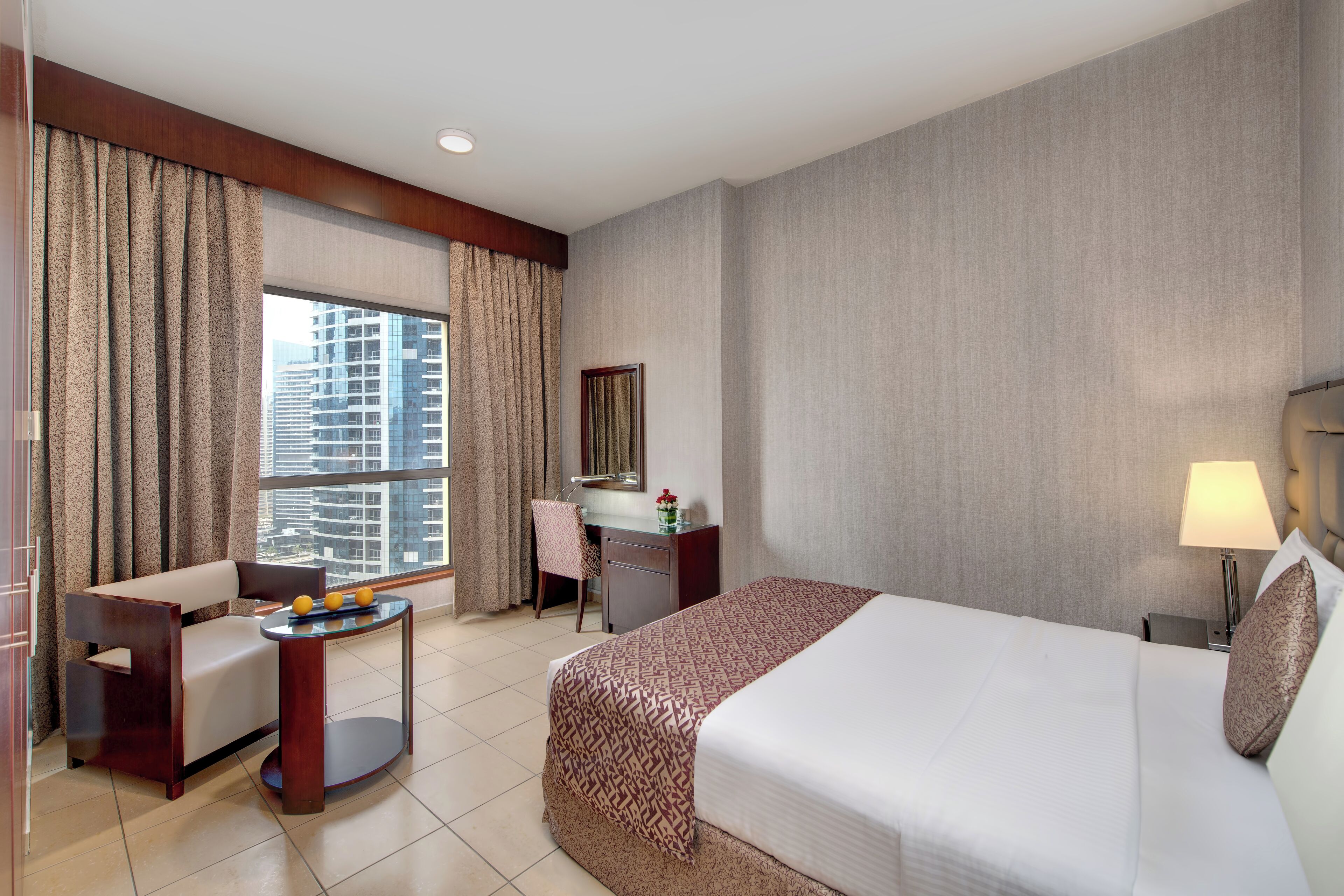 A hotel room with a bed, a desk, a chair, a small table with fruit, and a view of the city.