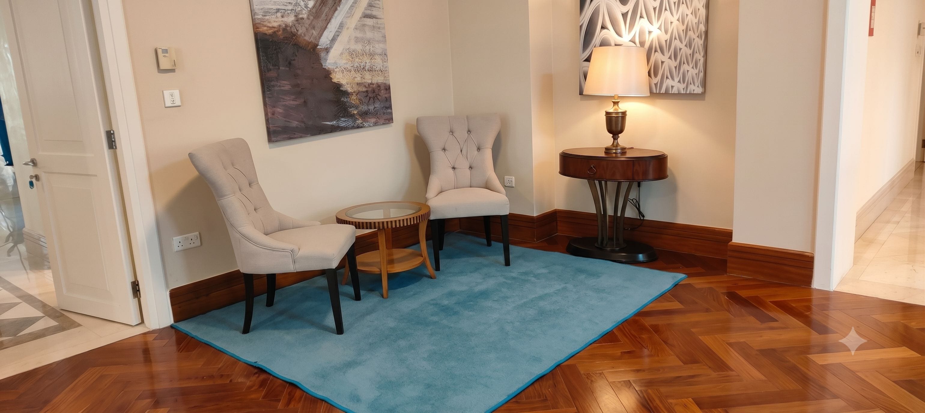 A room with two upholstered chairs, a small round table, a blue rug, a floor lamp, and a painting on the wall.