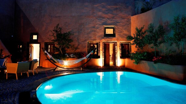 A pool area with a hammock, chairs, and a building with doors and windows. - Ducato Wine Village (Santorini)