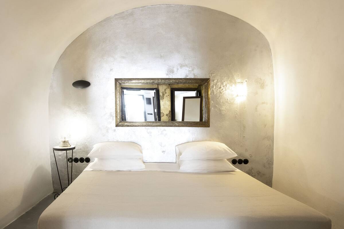A bedroom with a large bed, a small bedside table, a wall-mounted mirror, and a light fixture.