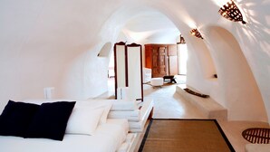 A modern, white-themed bedroom with a large bed, a small seating area, and a view of a wooden cabinet and a chair in the background. - Ducato Wine Village (Santorini)