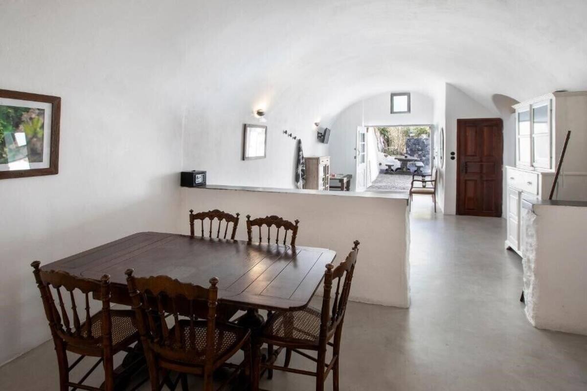 A dining area with a wooden table and chairs, a kitchen area with white cabinets, and a view of the outside.