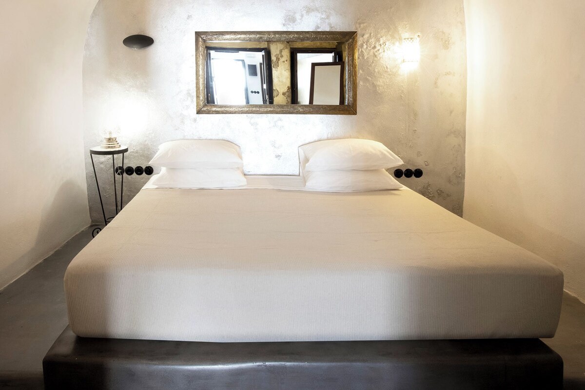 Un dormitorio con una cama grande, una mesita de noche pequeña, un espejo en la pared y una lámpara.