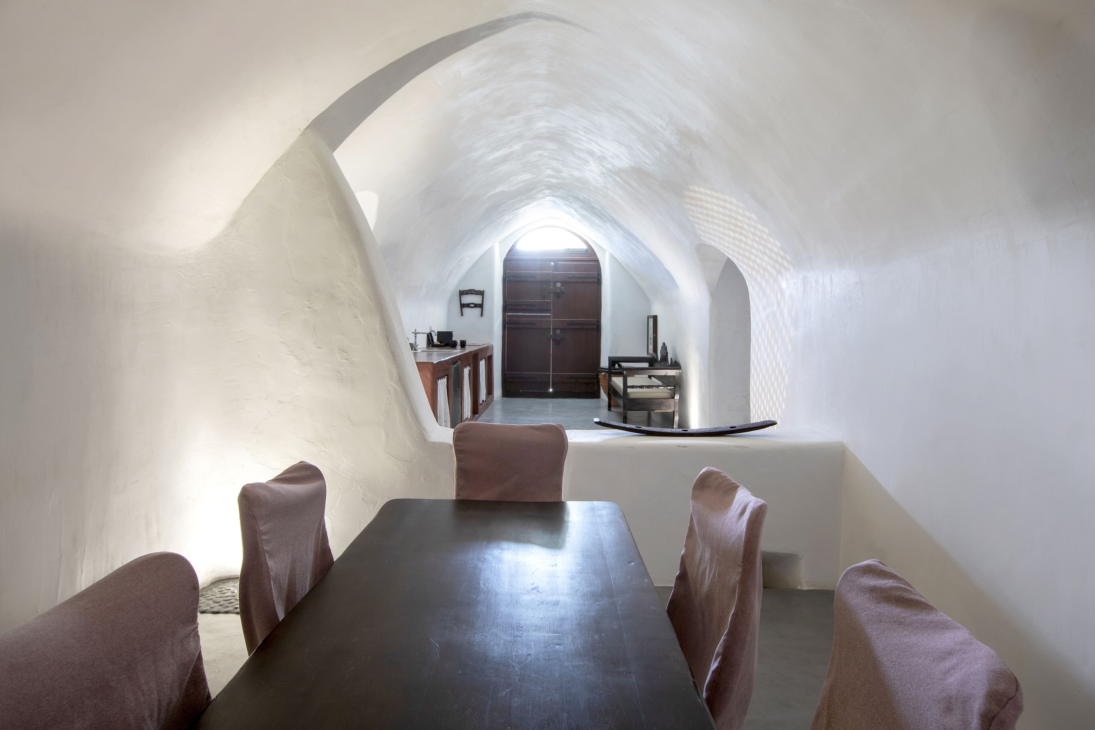 A dining area with a long table and chairs, an arched ceiling, and a view into a kitchen area.