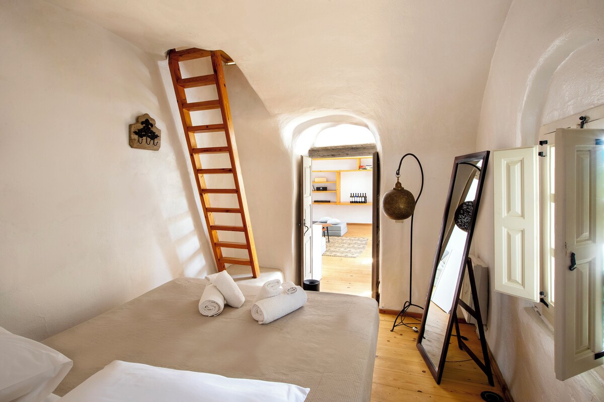 A bedroom with a bed, a ladder, a mirror, and an open door leading to another room.