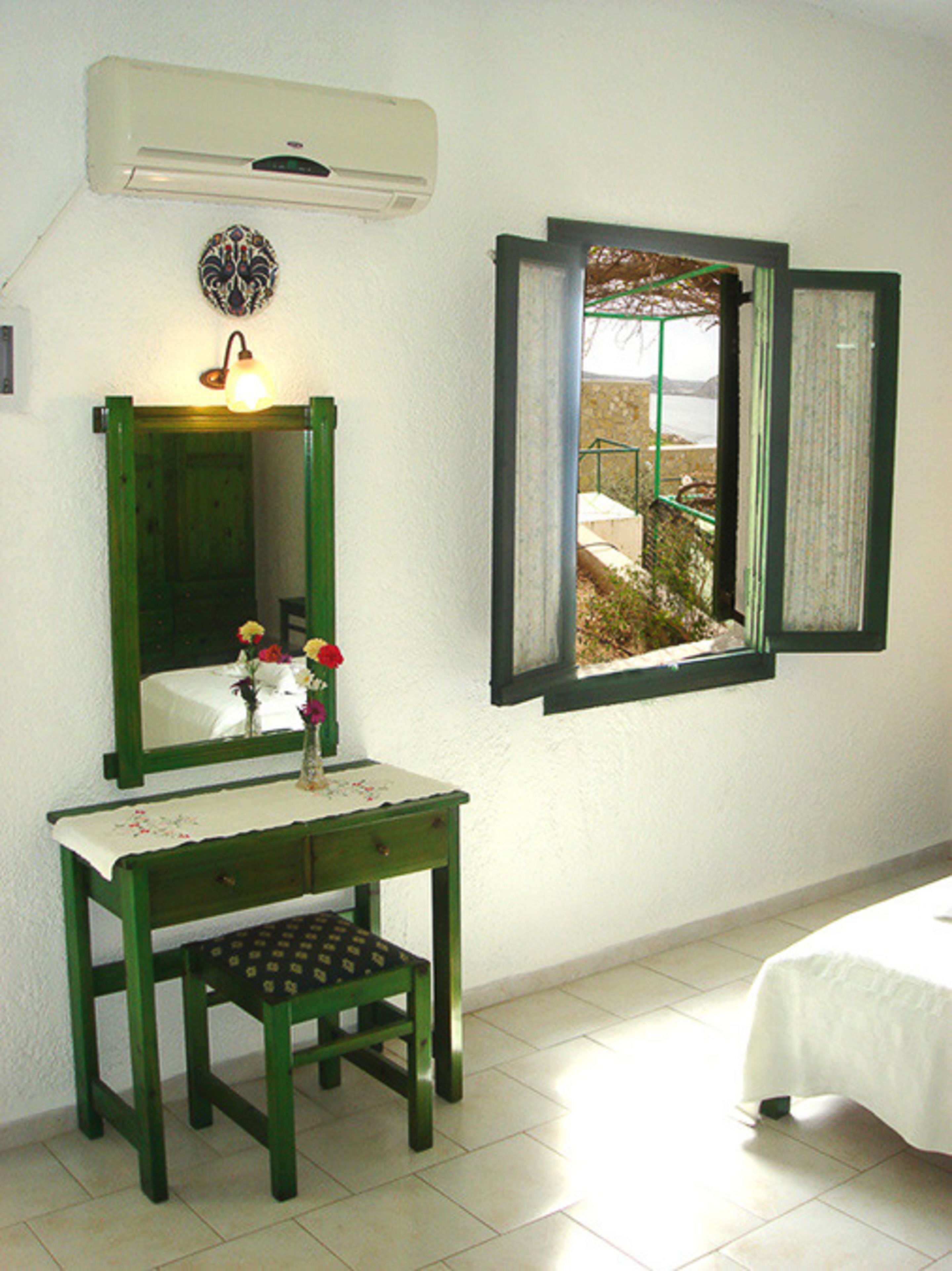 A room with a green vanity, a mirror, a small table with a vase of flowers, a bed, and a window with a view of the outdoors.