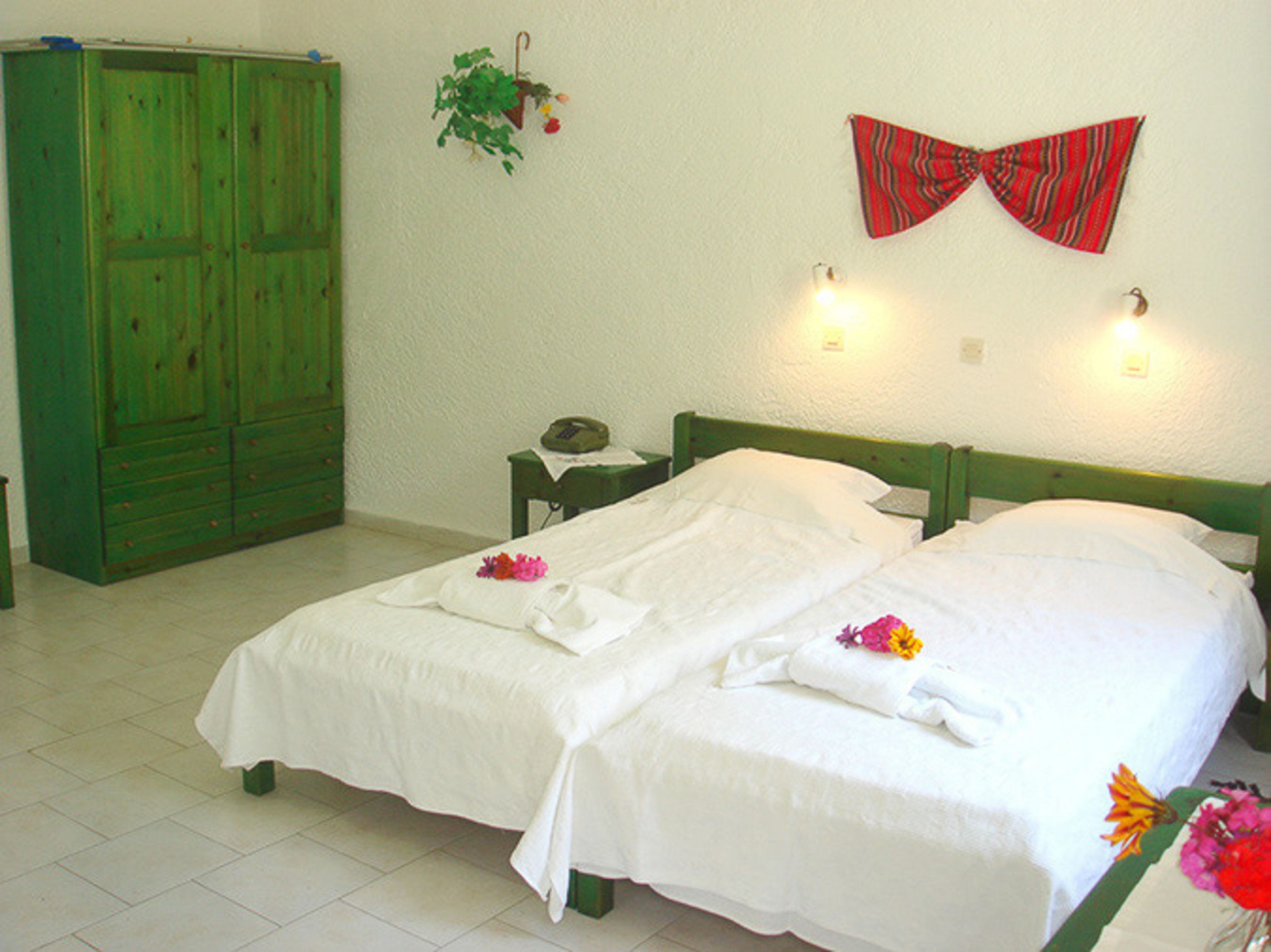 Een kamer met twee eenpersoonsbedden, een groene garderobe, een nachtkastje met een lamp, en een rode vlinderdas decoratie aan de muur.