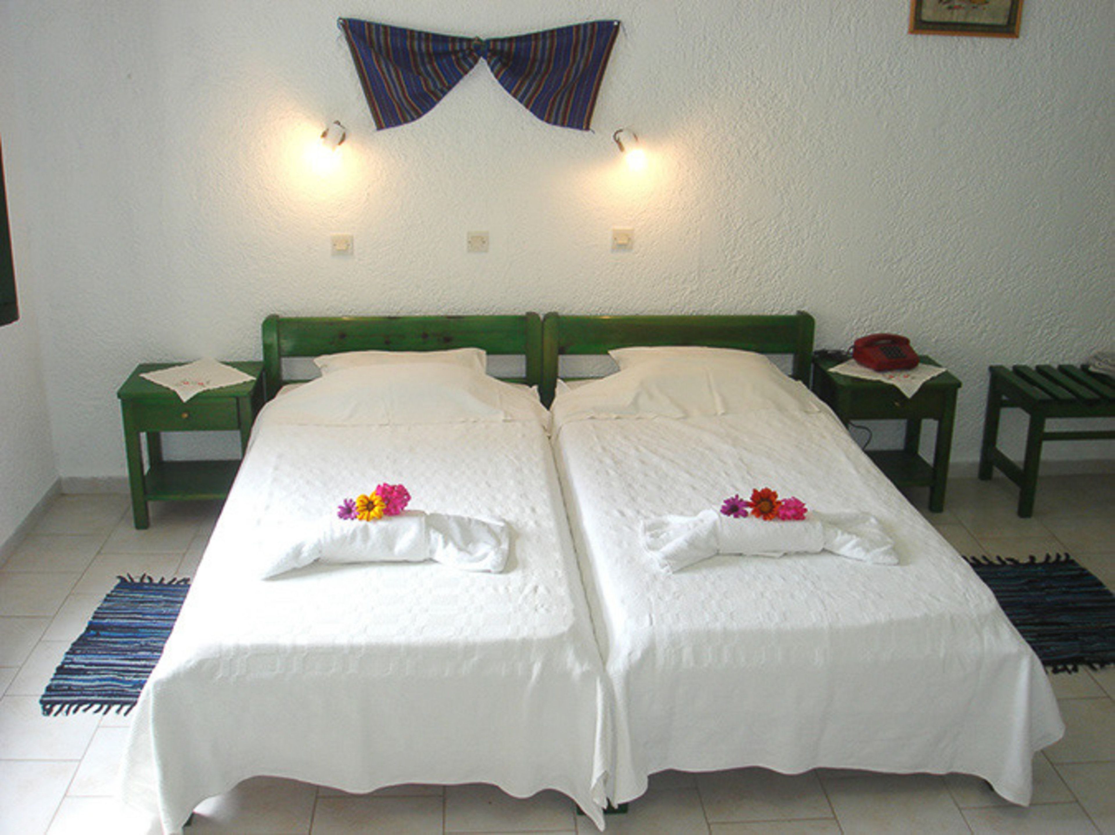 Un lit double avec du linge de lit blanc et des motifs floraux, deux tables de chevet, un fauteuil vert et un téléphone fixé au mur.