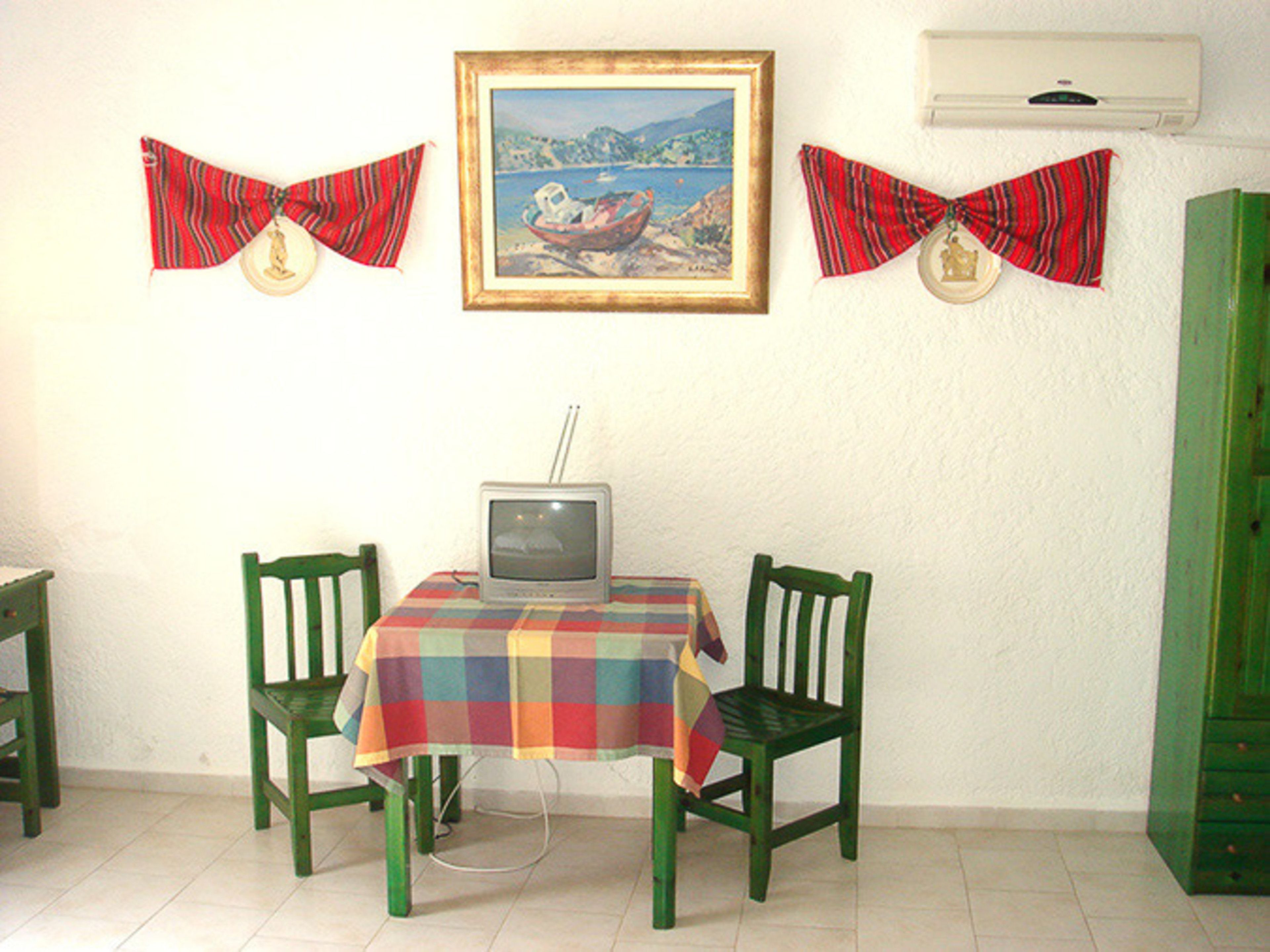 A dining area with a table covered by a multicolored tablecloth, two green chairs, a television on the table, and a framed painting above.
