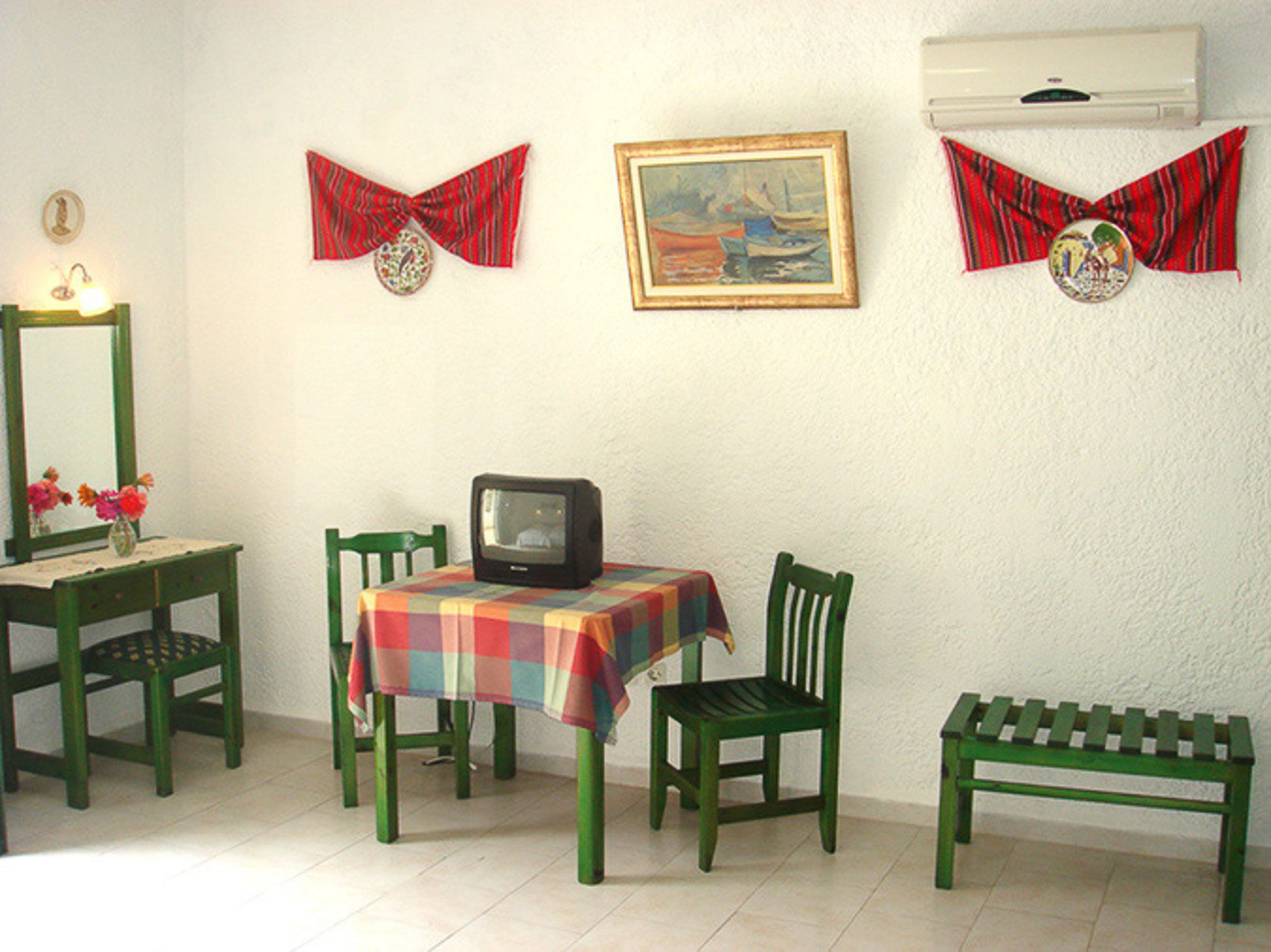 A room with a television on a table, two chairs, a bench, and wall decorations including a framed painting and two bow ties.