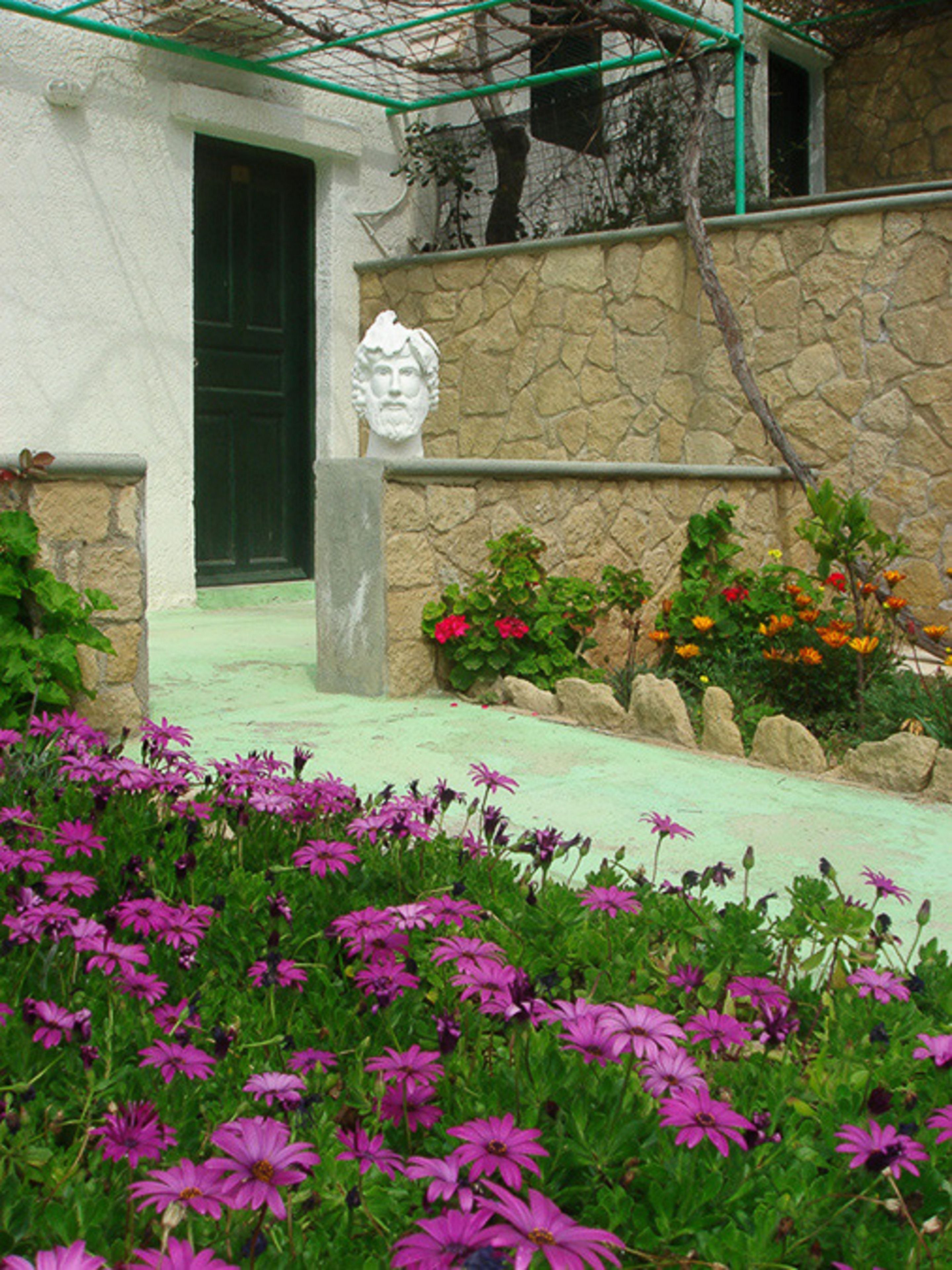 A stone wall with a bust sculpture, a green door, and a garden with purple flowers.