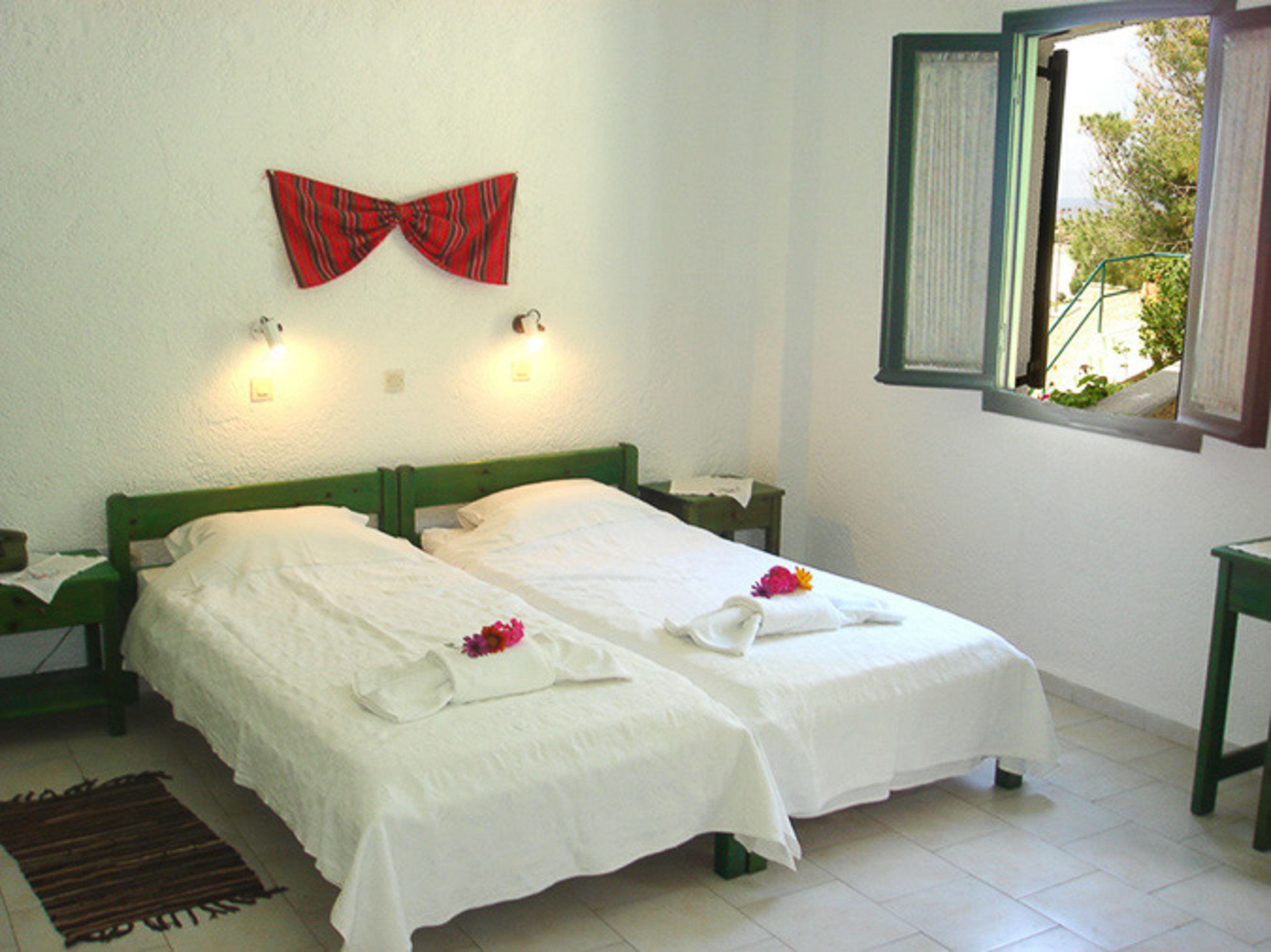 A room with two beds, a red bow tie on the wall, and a window with a view of greenery.