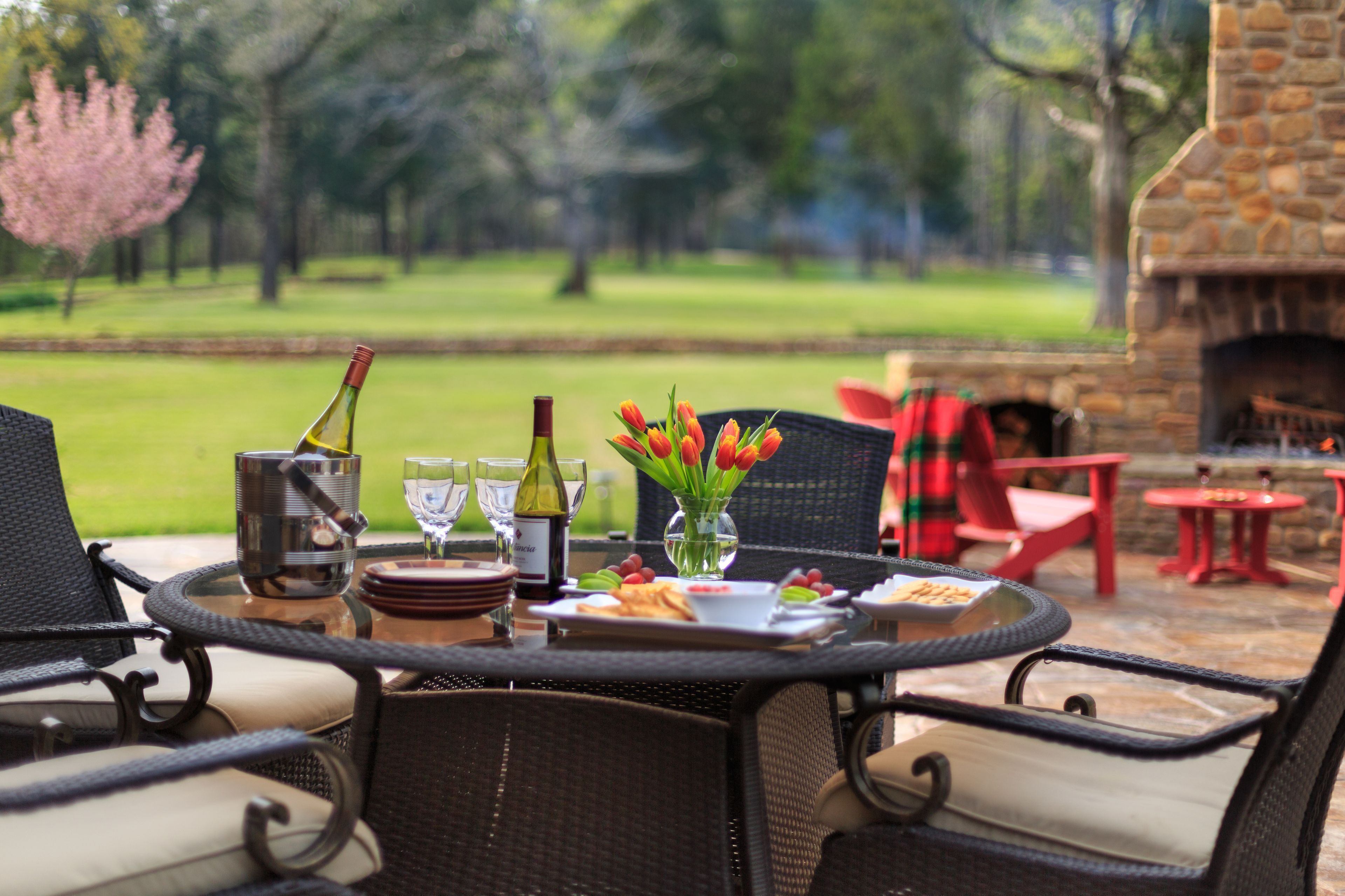 An outdoor dining area with wicker chairs, a stone fireplace, and a view of a grassy field.