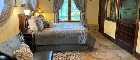 A bedroom with a large bed, a nightstand, a lamp, a window with curtains, a ceiling fan, and a door.