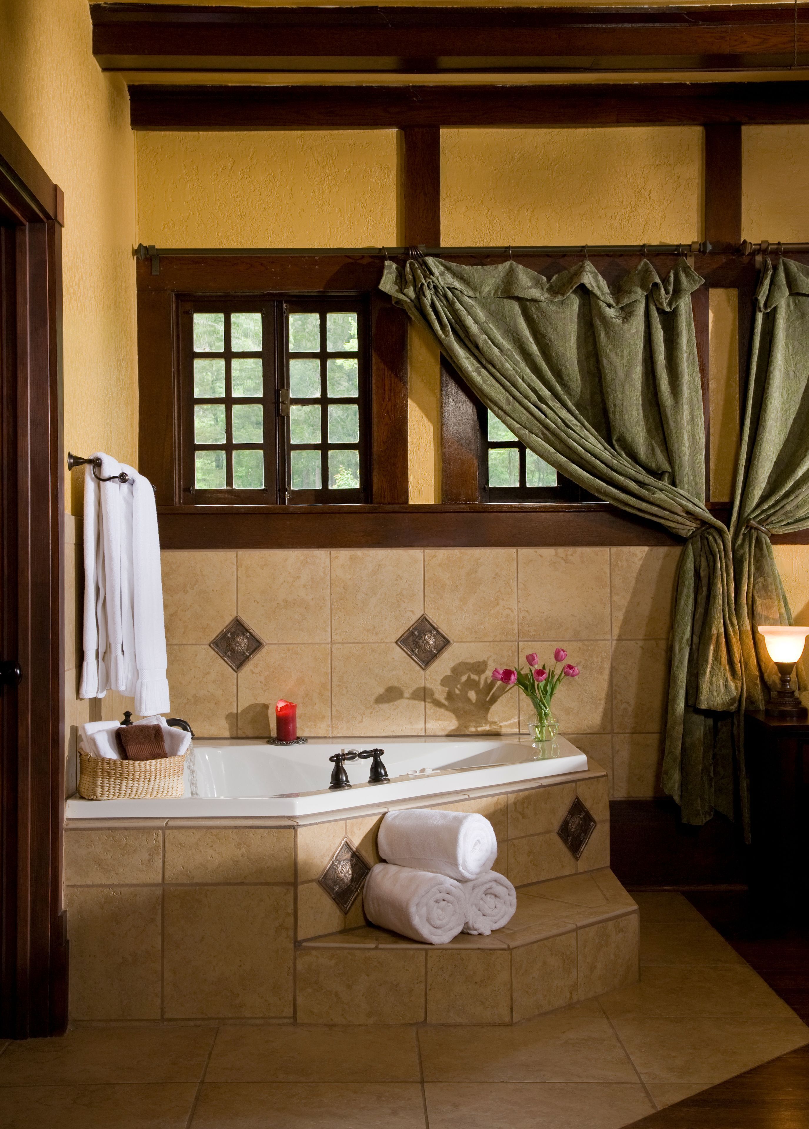A bathroom with a bathtub, a window with curtains, a vase with flowers, and a candle.