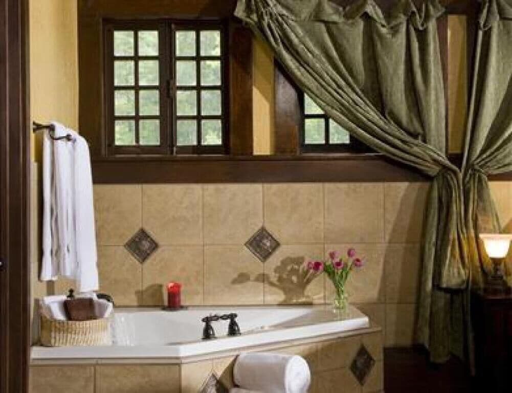 A bathroom with a bathtub, a window with curtains, a towel rack, a candle, and a vase with flowers.