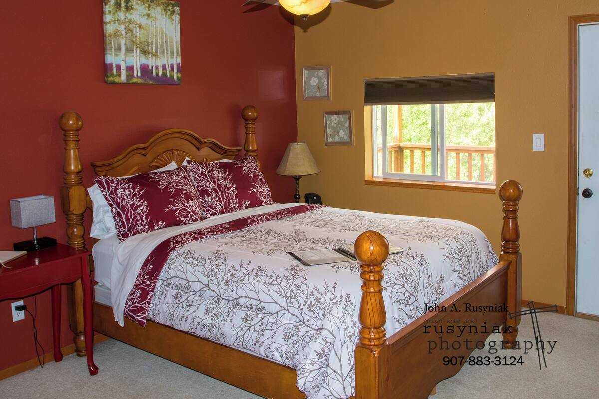 Quarto com cama de madeira, mesa de cabeceira, abajur, ventilador de teto, janela com vista e um quadro na parede.