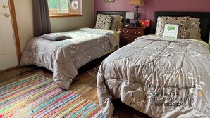 A bedroom with two beds, a window with blinds, a colorful rug, and a wall with framed pictures. - A Hyde Away Inn B&B (Tok)
