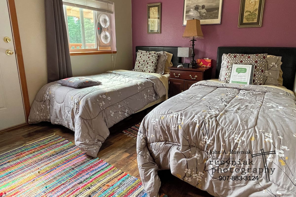 Quarto com duas camas, janela com persianas, tapete colorido e parede com quadros.