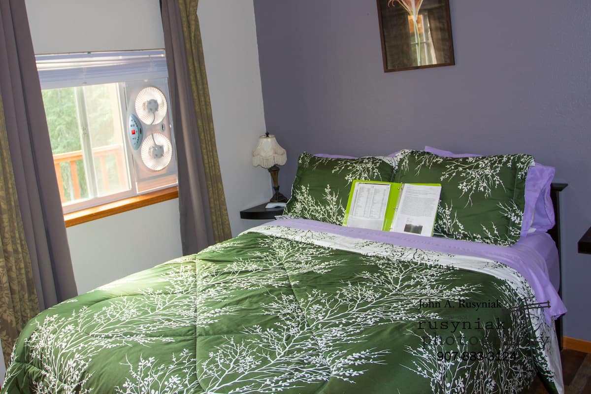 Quarto com cama, janela com cortinas, quadro na parede e mesa de cabeceira com abajur.