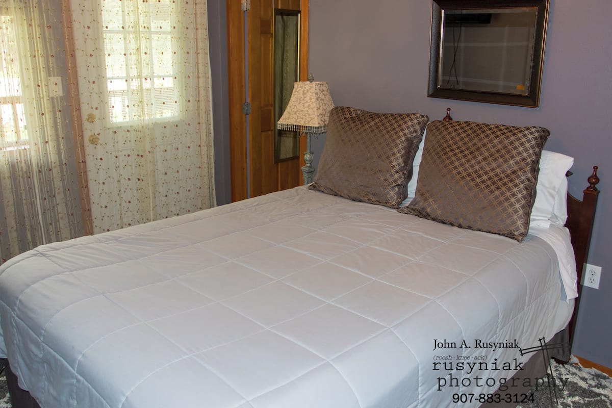Uma cama bem arrumada com um edredom branco e almofadas estampadas, uma cabeceira de madeira, um espelho fixado na parede e uma janela com cortinas florais.