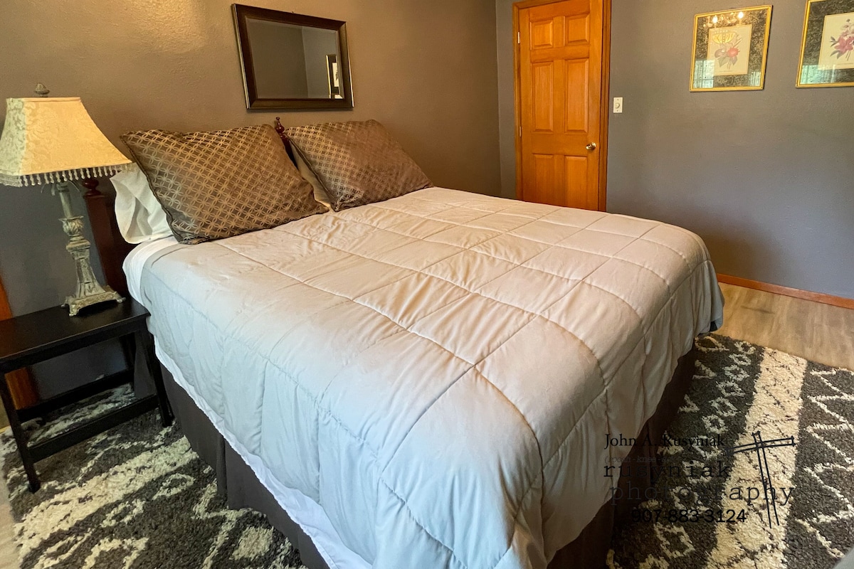 A bedroom with a bed, a nightstand, a lamp, a mirror, a door, and two framed pictures on the wall.