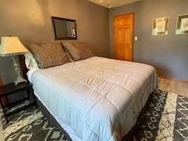 Un dormitorio con cama, mesita de noche, lámpara, espejo, puerta y dos cuadros en la pared.