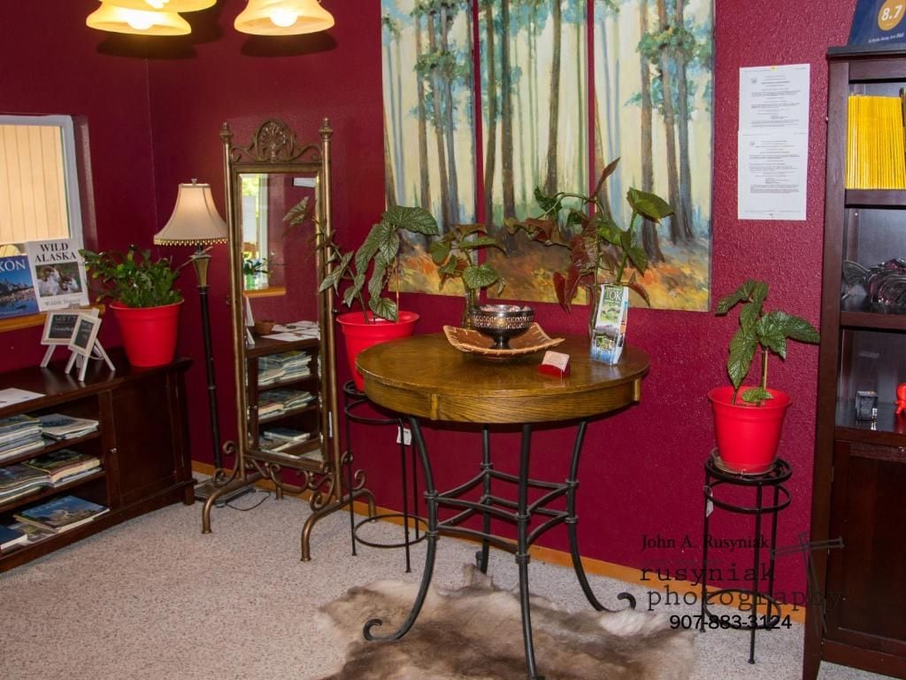 A room with a round wooden table, red potted plants, a bookshelf, a mirror, and a painting on the wall.
