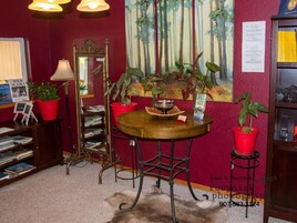 A room with a round wooden table, red potted plants, a bookshelf, a mirror, and a painting on the wall.