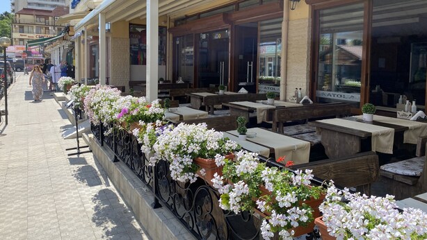 An outdoor cafe with wooden tables and chairs, potted plants, and a building with a sign in the background. - Hotel Atol (Sunny Beach)