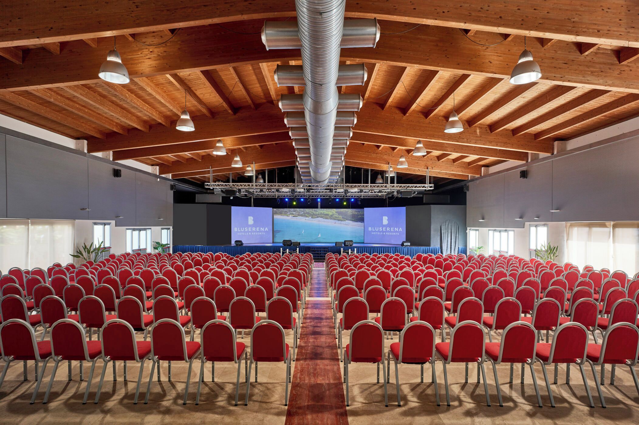 Una sala conferenze con file di sedie rosse, un palco con uno schermo grande e travi a cassettoni in legno.