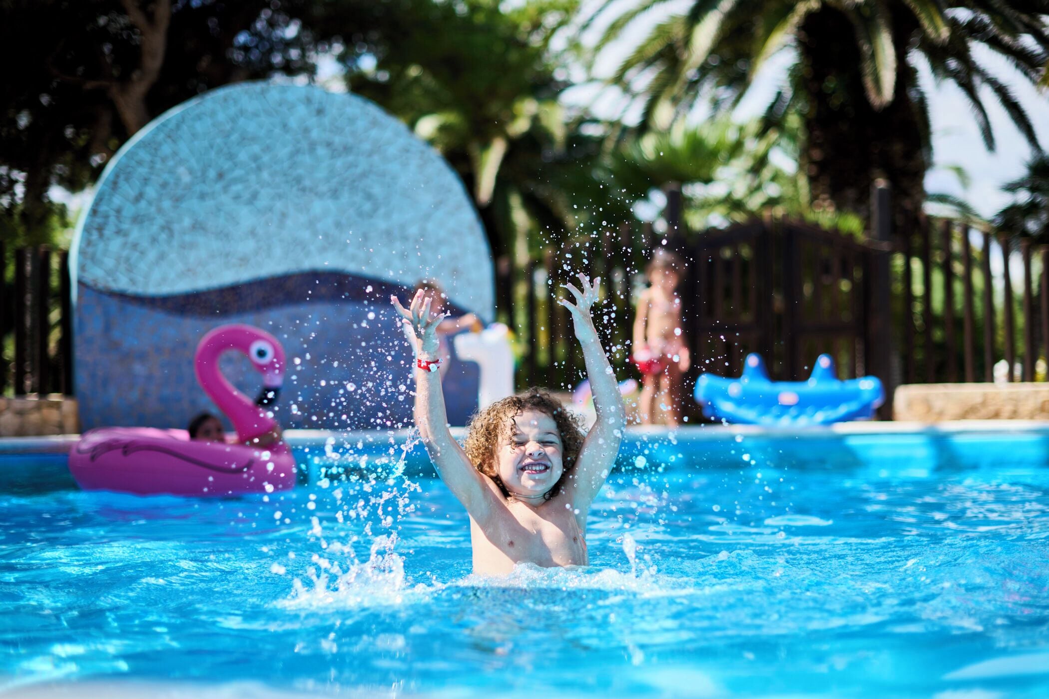 A swimming pool with a child playing, a pink flamingo float, and a large blue water feature in the background.