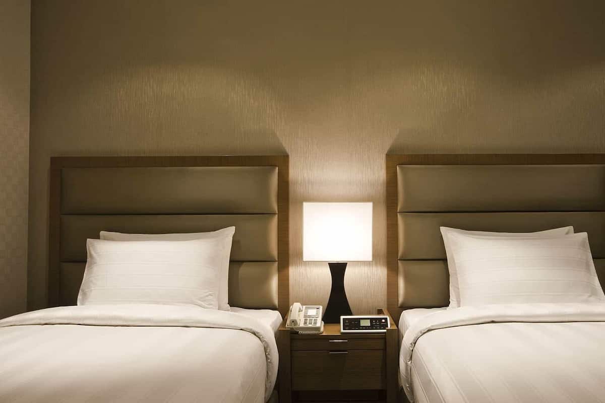 A hotel room with two beds, a bedside table with a lamp, and a phone.