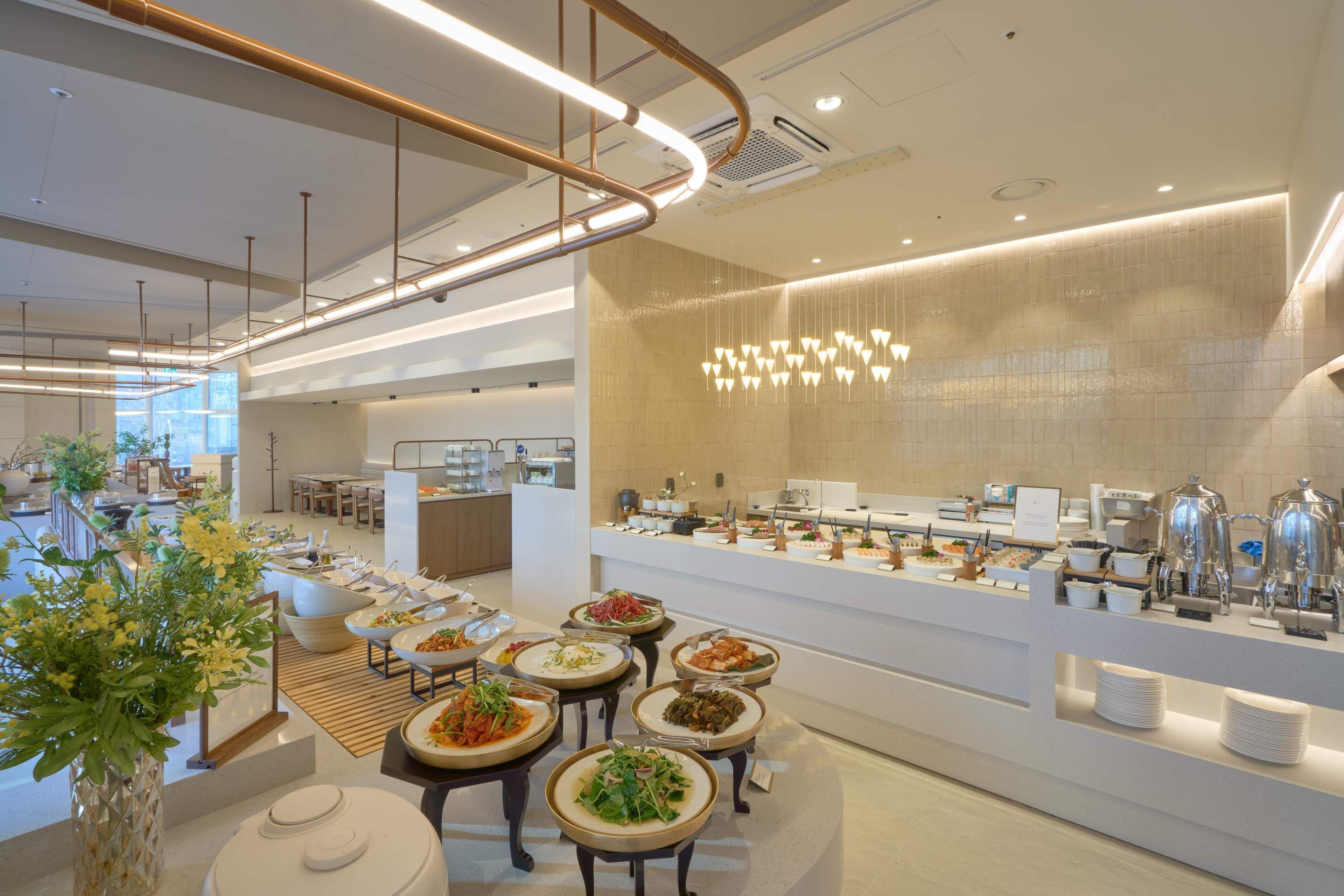Un comedor de hotel moderno con una barra de buffet, un área de estar con mesas y sillas, y una planta decorativa.