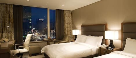 A hotel room with two beds, a desk with a lamp, a chair, and a view of the city at night.