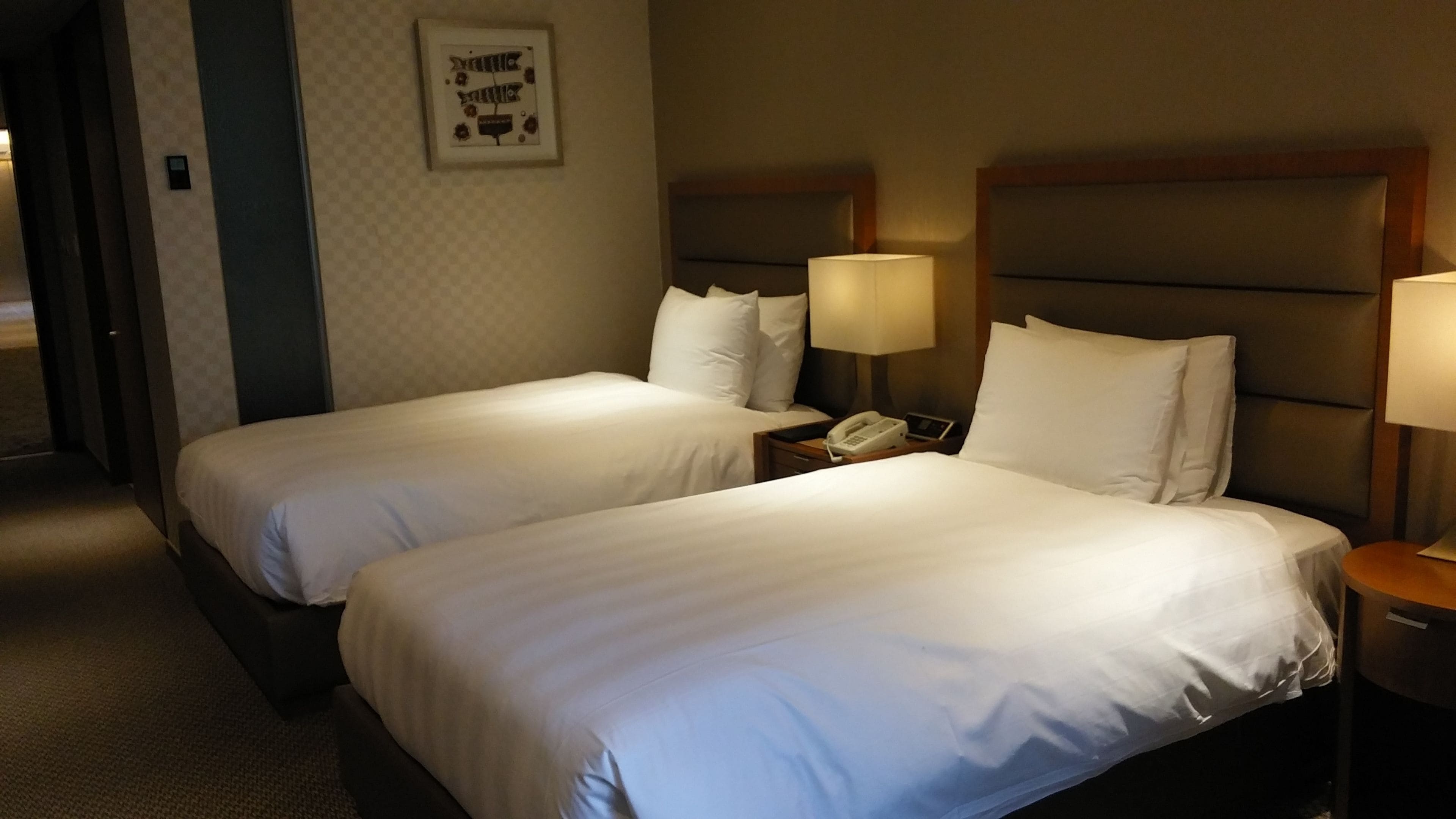 A hotel room with two single beds, each with a pillow and a bedside table with a lamp and a phone.