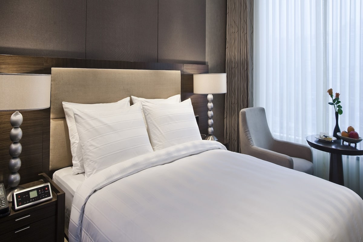 A modern hotel room with a large bed, a bedside table with a lamp, a chair, and a small table with a vase and fruit.