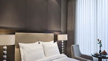 A modern hotel room with a large bed, a bedside table with a lamp, a chair, and a small table with a vase and fruit.