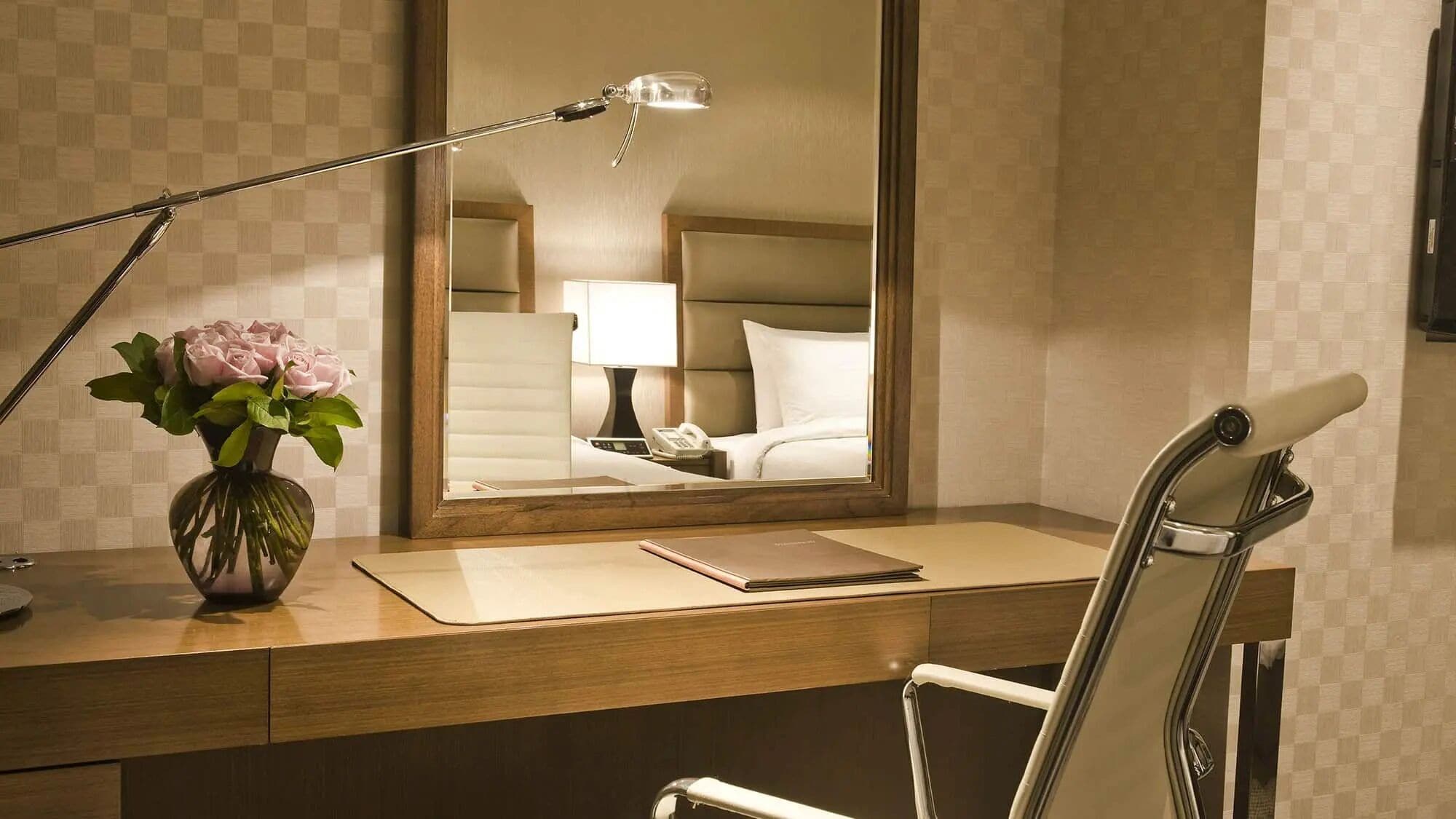 Habitación de hotel con escritorio, espejo, lámpara, un florero con flores y una silla.