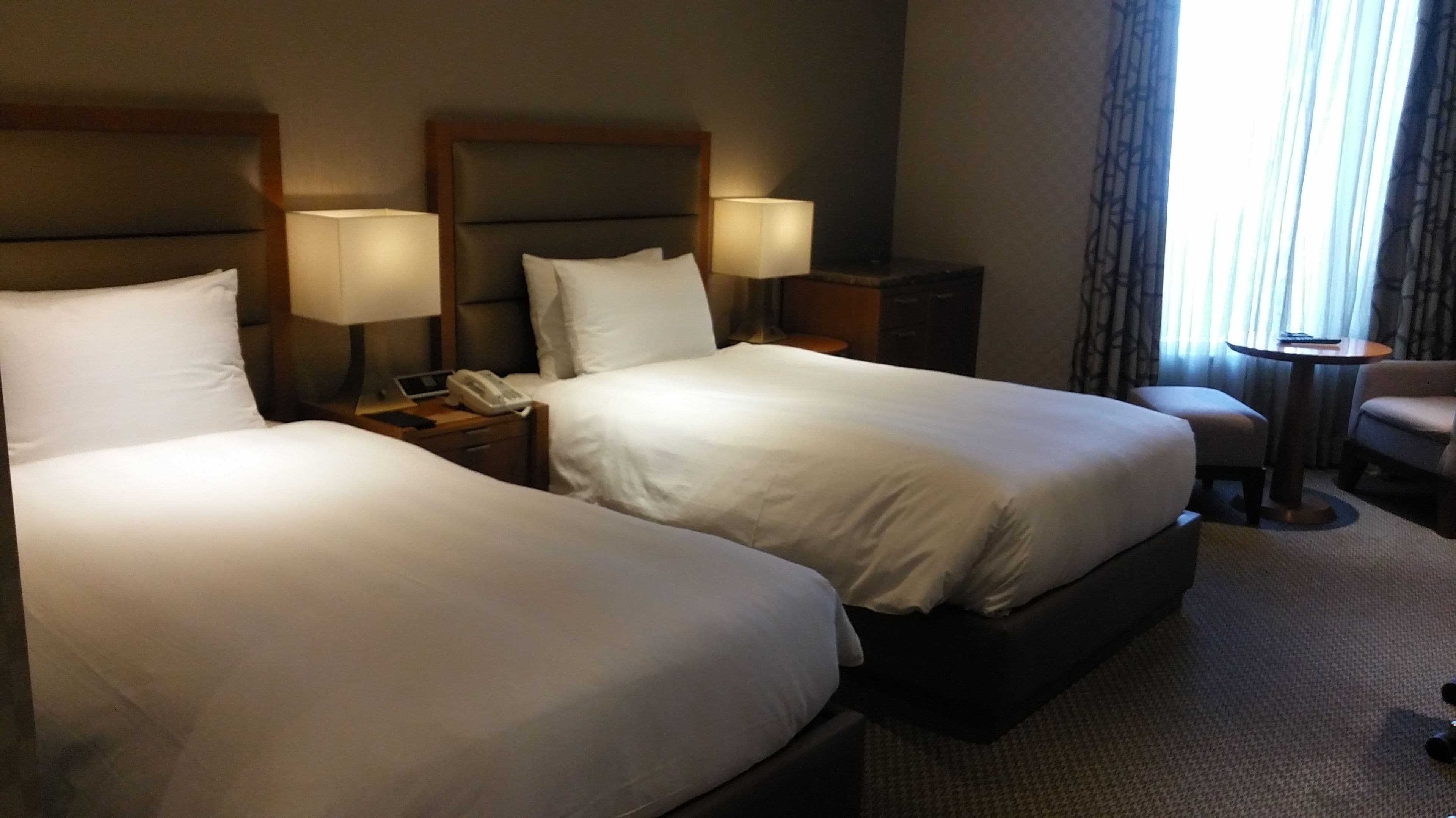 Habitación de hotel con dos camas, mesitas de noche con lámparas, una mesita pequeña y una silla.