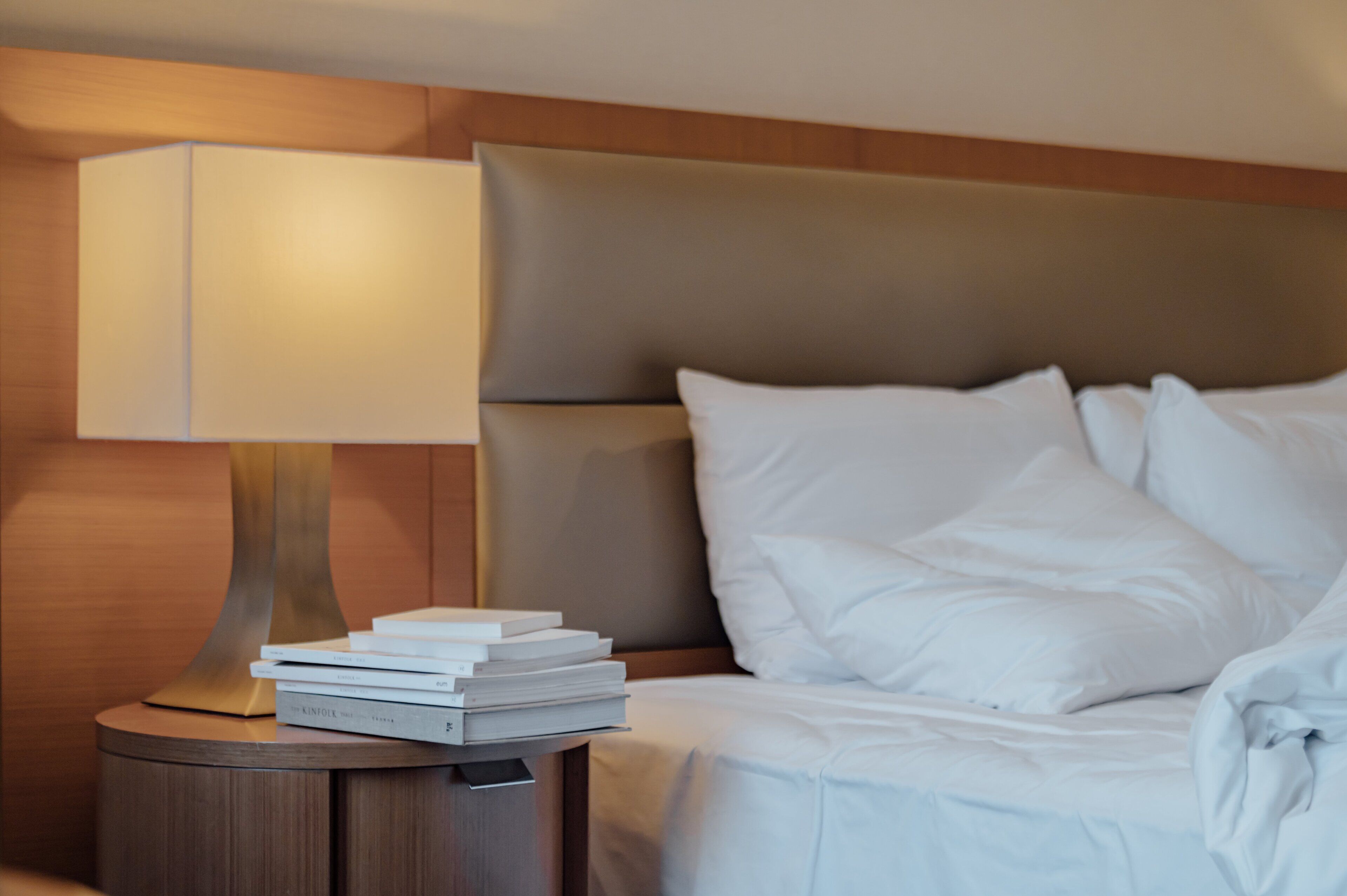 A neatly made bed with white linens, a bedside lamp, and a stack of books on a wooden nightstand.
