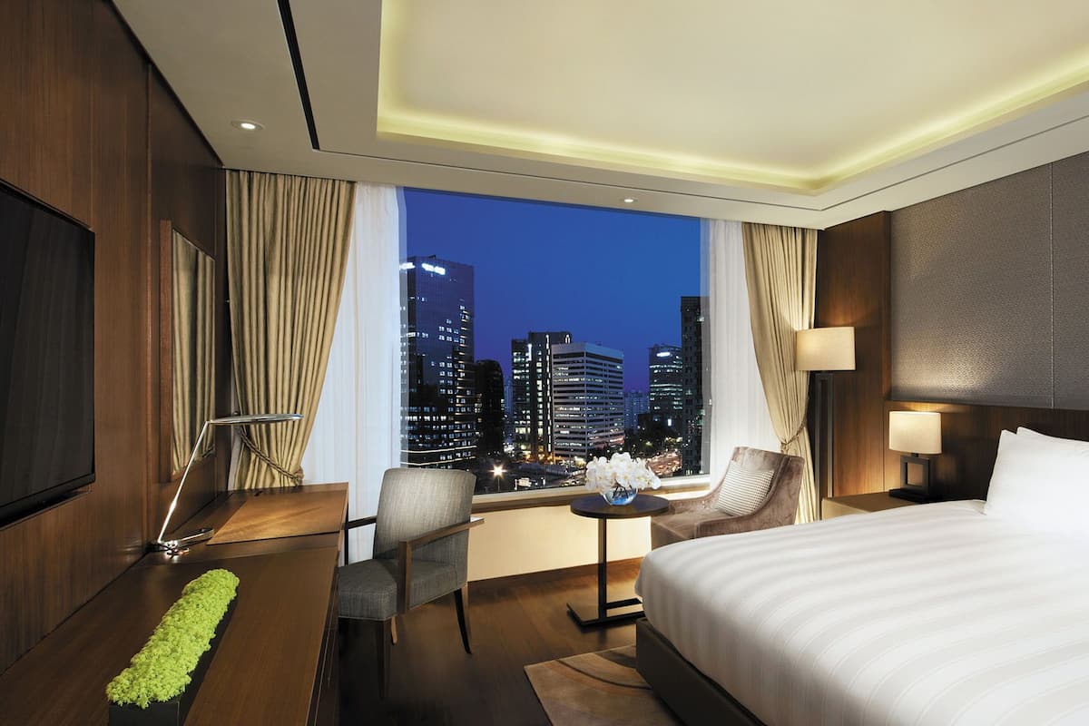 A hotel room with a large bed, a desk with a chair, a small table with a lamp, and a view of the cityscape at night.
