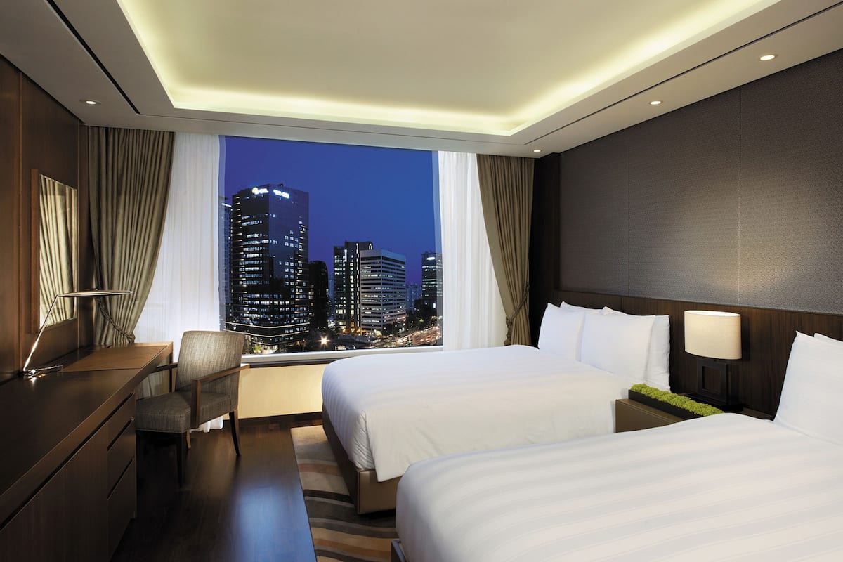 A hotel room with two beds, a desk, a chair, and a large window with a city view.